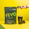 A person is pouring vanilla flavored protein powder into a shaker cup beside a green package labeled 'VIVO LIFE PERFORM PLANT PROTEIN'.